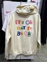 Canguro beige con capucha y cordones ajustables. Estampado en la espalda con la frase "it's ok not to be ok" en letras de colores y caritas sonrientes y tristes.