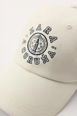 Gorra color crudo con visera y bordado frontal con el texto "Zara A Coruña Est. 1975 50 Years". Ajuste regulable en la parte posterior.