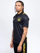 Remera polo de poliéster color negro con cuello clásico y tapeta de dos botones. Presenta el escudo del Club Atlético Peñarol bordado en el pecho y el año de fundación (1891) estampado en blanco. Tiene detalles en amarillo en los hombros y el escudo del club repetido en la parte inferior.