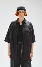 Camisa negra de manga corta, corte oversize, confeccionada en poplin 100% algodón. Presenta dos bolsillos delanteros, uno con solapa y otro con ojal y botón.