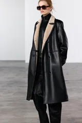 Abrigo largo de Zara Woman Collection, confeccionado en tejido efecto piel color negro. Tiene cuello solapa, manga larga, bolsillos laterales ocultos y forro interior de borreguillo color beige. El cierre es cruzado con botones.