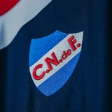 Camiseta de fútbol Umbro del Club Nacional de Football, modelo Away Identity 2024. Diseño azul con detalles en rojo y blanco en cuello y mangas. Incluye logo de Umbro, escudo del club y sponsor Antel en el frente.