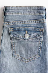 Jeans de denim azul claro con efecto lavado y desgaste sutil. Tienen un corte ajustado hasta la rodilla y luego se abren en un estilo acampanado (flare). Son de tiro bajo y presentan bolsillos delanteros abiertos y bolsillos traseros con solapa y botón.