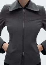 Chaqueta gris con finas rayas verticales, corte entallado, cuello camisero, cierre frontal con cremallera doble, bolsillo de ribete en el pecho y mangas largas.