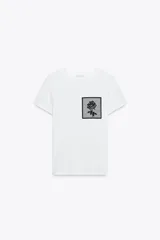 Camiseta blanca de algodón con cuello redondo y mangas cortas. Presenta un bolsillo de tul con una flor bordada y abalorios en el delantero.