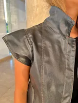 Chaleco sin mangas de gabardina con acabado foil (efecto satinado/brillante), color gris oscuro o negro. Presenta cuello alto y pespuntes contrastantes visibles en el frente y hombros.