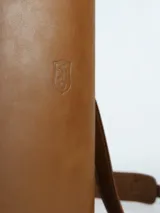 Matera cilíndrica de cuero color camel, con cierre superior y correa ajustable para llevar al hombro. Presenta un escudo grabado en el frente.