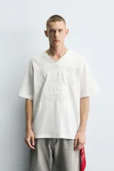 Camiseta blanca de corte holgado con cuello en V y mangas cortas. Presenta un diseño de malla con diferentes texturas combinadas a tono y estampado frontal con la inscripción "Legacy Crew 95 Elite Mode".