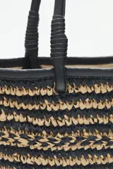 Bolso tote tejido en raffia color negro y beige, con vivos y correas en cuero sintético negro. Tiene cierre interno con snap metálico y forro de algodón.