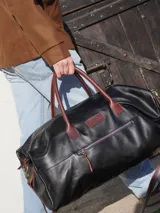 Bolso de viaje tipo weekender de cuero color marrón con detalles en rojo en los cierres. Tiene doble asa corta y correa de hombro ajustable y extraíble. Cuenta con bolsillos internos y externos.