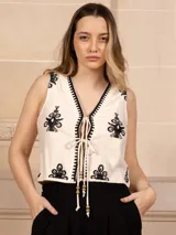 Chaleco corto color crudo con bordados florales negros, escote en V con borde negro y lazos de ajuste con cuentas de madera.
