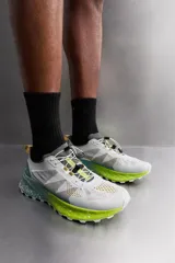 Zapatilla deportiva para carreras de montaña, color blanco con detalles en gris, verde y amarillo. Parte superior fabricada en tejido de punto transpirable con zona de puntera y talón recubiertas de material Shield Protect Max que protege de la abrasión. Ajuste de los cordones mediante pieza técnica. Tirador en la parte posterior para facilitar el calce. Plantilla interior OrthoLite® extraíble. Suela con tecnología Spume Foam que garantiza ligereza y confort.
