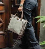 Cartera tipo tote de cuero color gris claro con detalles en marrón. Tiene doble asa corta y flecos decorativos en los laterales.