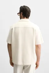 Camisa color crudo de corte regular fit, confeccionada en tejido con textura. Tiene cuello bowling, manga corta, bajo con aberturas laterales y cierre frontal con botones.