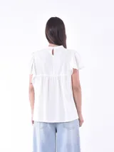 Blusa blanca de manga corta con volados, cuello alto y detalles de encaje o bordado vertical en el frente. Presenta un corte holgado con frunce bajo el busto.