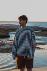 Buzo de manga larga reversible. Un lado es a rayas horizontales azul marino y blanco, con un logo rojo bordado en el pecho y un parche rojo en la manga. El otro lado es de color azul claro liso, con un cuello redondo deshilachado.