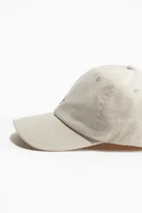Gorra color crema de sarga de algodón con diseño bordado de un pájaro en el frente. Tiene ojales bordados en la parte superior, trabilla ajustable con hebilla de metal atrás y cinta antitranspirante de algodón.