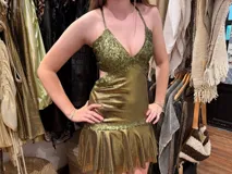 Vestido corto dorado con escote en V, cut outs laterales bajo el busto, breteles finos que se cruzan en la espalda y atan atrás, busto, pieza bajo busto y cinto cadera en tela combinada y dos volados en el ruedo.