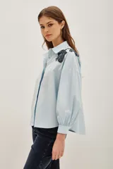 Camisa blanca de algodón con cuello clásico, mangas largas abullonadas con puños anchos y cierre frontal de botones. Presenta aplicaciones bordadas en dorado con detalles de animal print en los hombros.