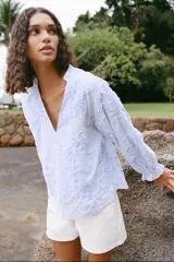 Blusa celeste con rayas verticales blancas, cuello redondo con escote en pico y mangas largas abullonadas terminadas en puño elástico. Presenta detalle de tejido en relieve con forma de flor.