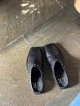 Mules de cuero negro con punta cuadrada y detalle de tachas plateadas en el borde superior.