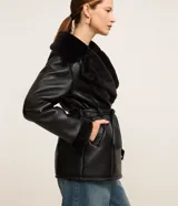 Blazer negro confeccionado en material sintético PU, con cuello y puños de piel sintética, cierre por cremallera y cinturón con hebilla en la cintura.