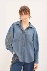 Camisa de denim azul de corte amplio, manga larga, cuello camisero y bolsillos en el delantero. Presenta bordados dorados de mostacillas, lentejuelas y canutillos en mangas y parte inferior de un lateral del pantalón.