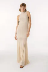Vestido largo color beige, con cuello alto y sin mangas. La parte superior es ajustada al cuerpo y la falda es amplia y fluida, con paneles traslúcidos.