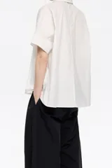 Camisa blanca de corte oversize confeccionada en 100% algodón, con cuello camisero, manga corta recta y ancha, cierre frontal con botones y pespuntes a contraste.