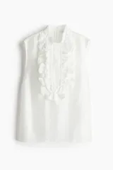 Blusa blanca sin mangas con cuello mao y pechera de volados con bordados de encaje.