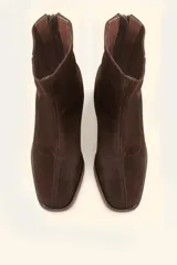 Bota corta de cuero vacuno color marrón oscuro, con diseño sin forro y taco cilíndrico de madera tallada a mano. Presenta cierre posterior y punta redondeada.