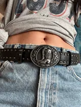 Cinturón negro de cuero sintético con hebilla plateada con diseño de botas cowboy y tachas plateadas.