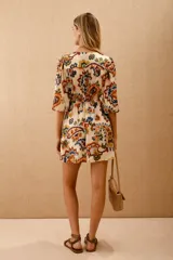 Vestido corto estampado con motivos florales en tonos naranja, azul y verde sobre fondo blanco. Tiene mangas 3/4 con volados en los puños, escote en V y cintura elastizada con lazo.