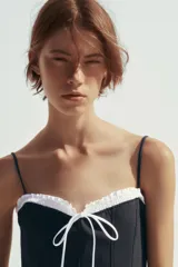 Vestido corto negro con tirantes finos ajustables, escote recto con volantes blancos y lazo decorativo. Cierre lateral con cremallera oculta.