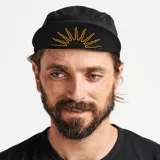 Gorro tipo camper negro con textura microperforada, visera corta y construcción plegable. Presenta un bordado frontal de un sol estilizado en tonos naranjas y un bordado lateral de una serpiente con una hoja en tonos amarillos y blancos.