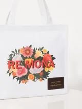 Bolso tipo tote de algodón blanco con estampado de flores y texto "RE MONA" en rojo. Tiene asas de mano y un parche de cuero sintético con texto grabado.