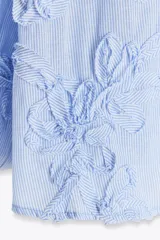 Blusa celeste con rayas verticales blancas, cuello redondo con escote en pico y mangas largas abullonadas terminadas en puño elástico. Presenta detalle de tejido en relieve con forma de flor.