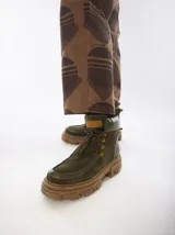 Botas tobilleras de cuero color verde oliva con suela de plataforma gruesa color marrón. Presentan puntera y detalles en material sintético brillante, cuello acolchado y cordones marrones con ojales dorados. Tienen una lengüeta trasera para facilitar el calce.
