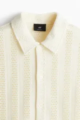 Camisa de manga corta color crema, confeccionada en tejido de punto jacquard con textura calada. Tiene cuello inglés, tapeta clásica con botones y bajo recto.