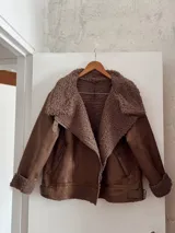 Campera marrón de gamuza sintética con cuello, puños y ruedo de piel sintética color marrón claro. Tiene cierre cruzado con cremallera y bolsillos laterales con cremallera.