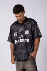 Camiseta de fútbol negra con estampado abstracto en gris, cuello tipo chomba y logos estampados en el frente.