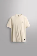 Camiseta color crema de corte relaxed fit, cuello redondo y manga corta. Tiene un texto estampado en el frente y un logo estampado en el ruedo.