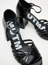 Sandalias negras de charol con tiras cruzadas en el empeine, pulsera con hebilla al tobillo y tacón cuadrado.