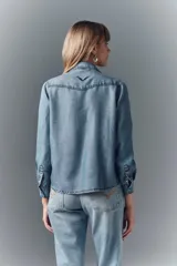 Camisa de denim celeste con apliques de piedras plateadas en los hombros, corte recto, cuello camisero, botones metálicos y bordado Rapsodia en la espalda.