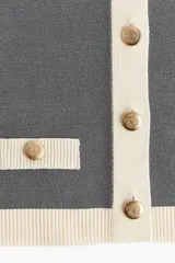 Cárdigan corto gris de punto fino con cuello redondo, cierre frontal con botones metálicos dorados y bolsillos decorativos con botón.