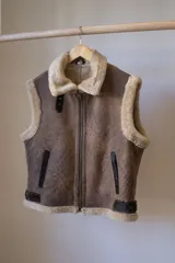 Chaleco marrón de cuero genuino con cuello y borde inferior de piel sintética color crema. Cierre frontal con cremallera metálica.