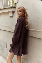 Vestido corto color marrón oscuro, estilo camisero, con cuello mao con ribete de volado y tapeta de botones con el mismo detalle. Posee mangas largas con puños sencillos de volado y un cinturón fino extraíble para ajustar la cintura. La falda tiene un fruncido escalonado que le da vuelo.