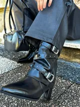Botas de cuero sintético negro con punta fina, caña media con correas y hebillas plateadas, y taco cuadrado.
