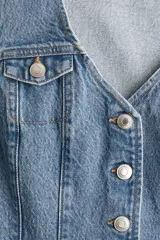 Chaleco ajustado de denim azul claro, sin mangas, con escote en V profundo y botonadura frontal de remaches metálicos. Presenta bolsillos de parche con solapa en el pecho y costuras verticales que definen la silueta.