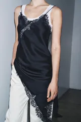 Vestido satinado negro con escote pico y tirantes finos. Presenta aplicación de encaje en el escote y en el bajo asimétrico, que incluye un tajo o caída pronunciada.